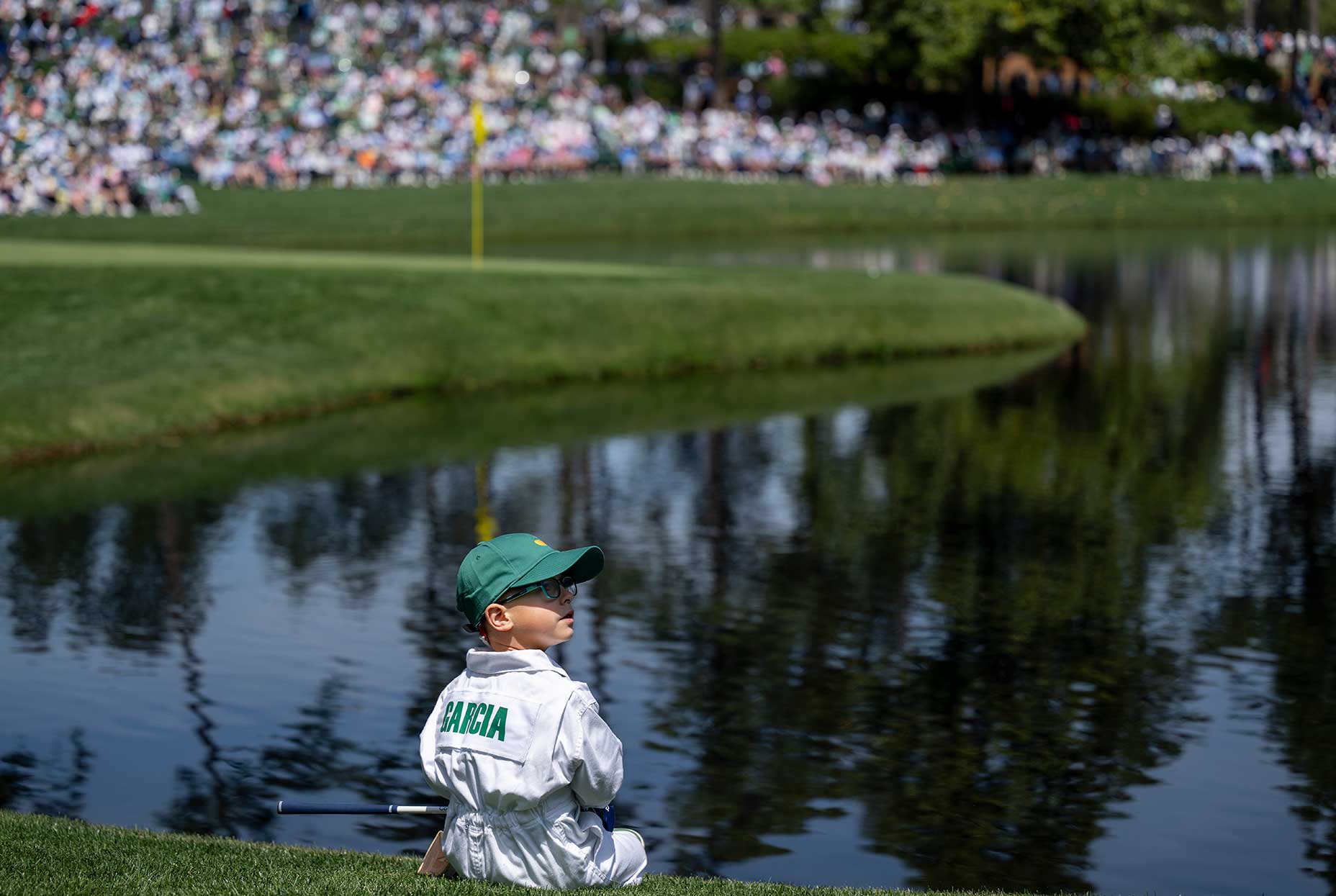 A young boy in a white caddie jumpsuit and green cap sits by a pond on the golf course at the Masters par 3 contest, with spectators and a yellow flag in the background. Garcia is printed on the back of his suit.