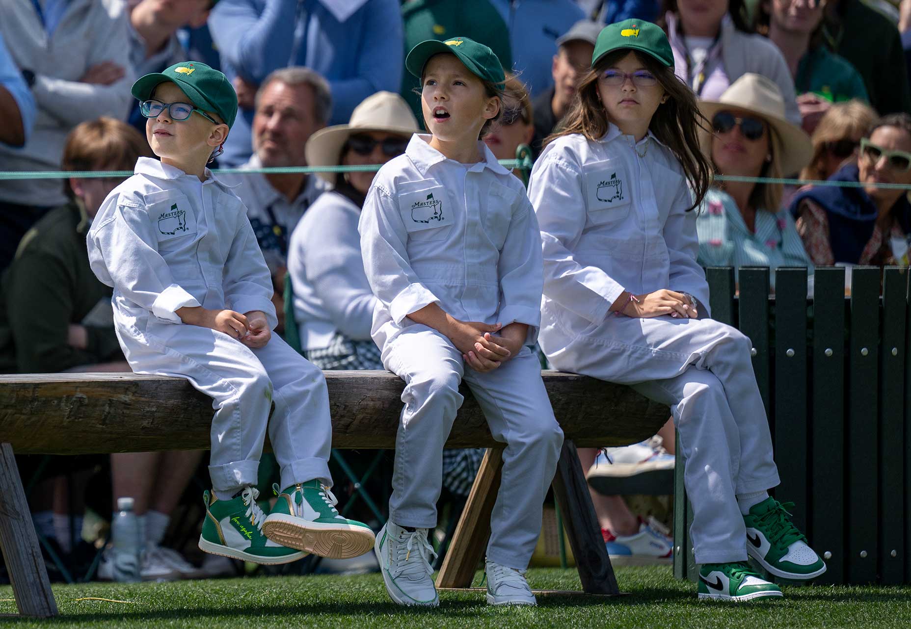 Three children in white jumpsuits and green hats, reminiscent of the Masters Par 3 tradition, sit on a wooden bench outdoors, watching something intently amid spectators in sunglasses and hats on a sunny day.