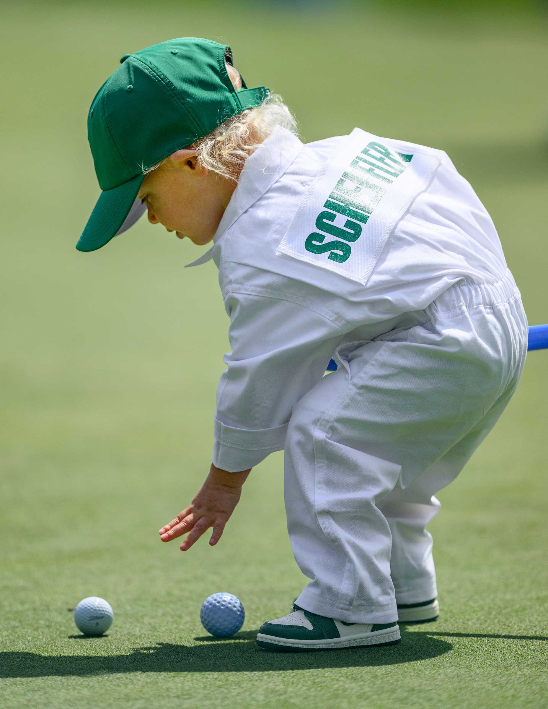 A young child dressed in a white outfit and green cap bends down to touch a golf ball on the green during the Masters par 3 event, with a name badge reading “SCHUFFELE” on their back. Two golf balls are on the ground in front of them.