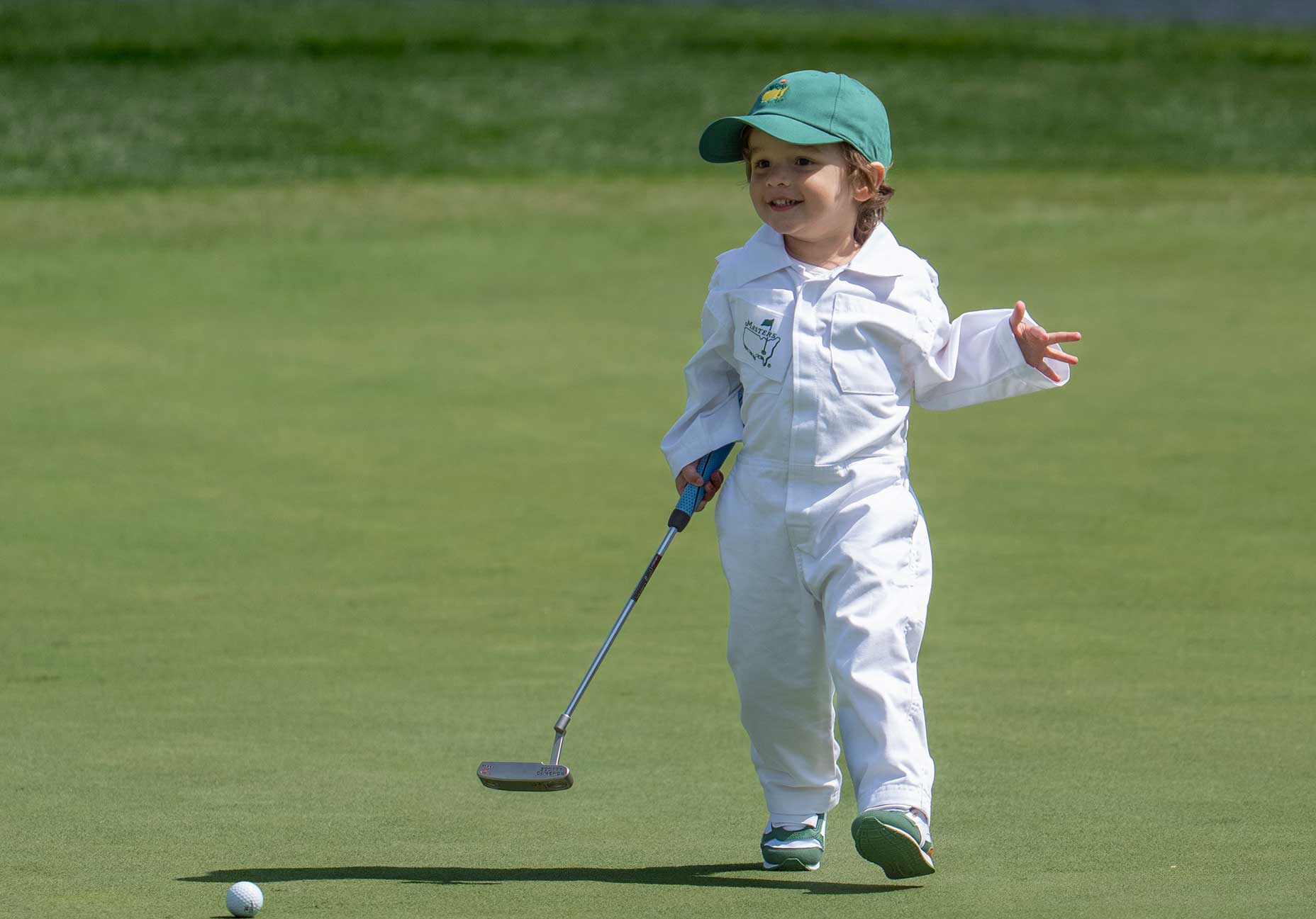 A young child in a white jumpsuit and green cap smiles while holding a golf putter on a Masters Par 3 green, standing near a golf ball.