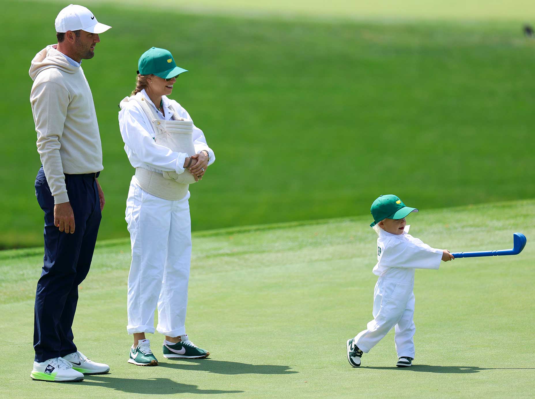 A man, a woman holding a baby in a carrier, and a young child with a toy golf club stand on a golf course. The child, dressed in white with a green cap, looks ready for the Masters as he swings on the par 3.