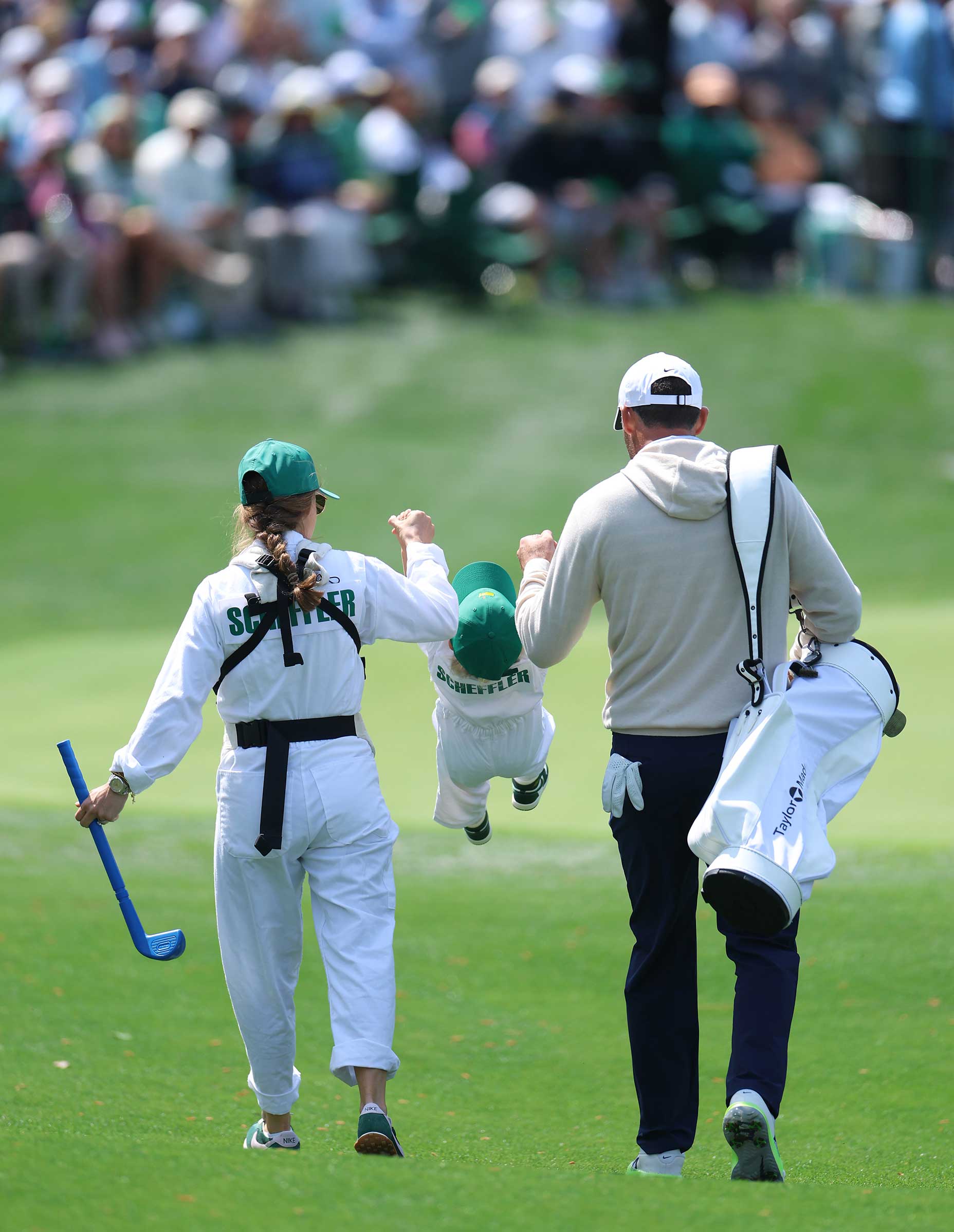 A woman and a man in golf attire swing a child joyfully between them as they walk on a lush par 3 hole. The crowd in the background watches the scene under a bright sky, capturing the family spirit of the Masters.