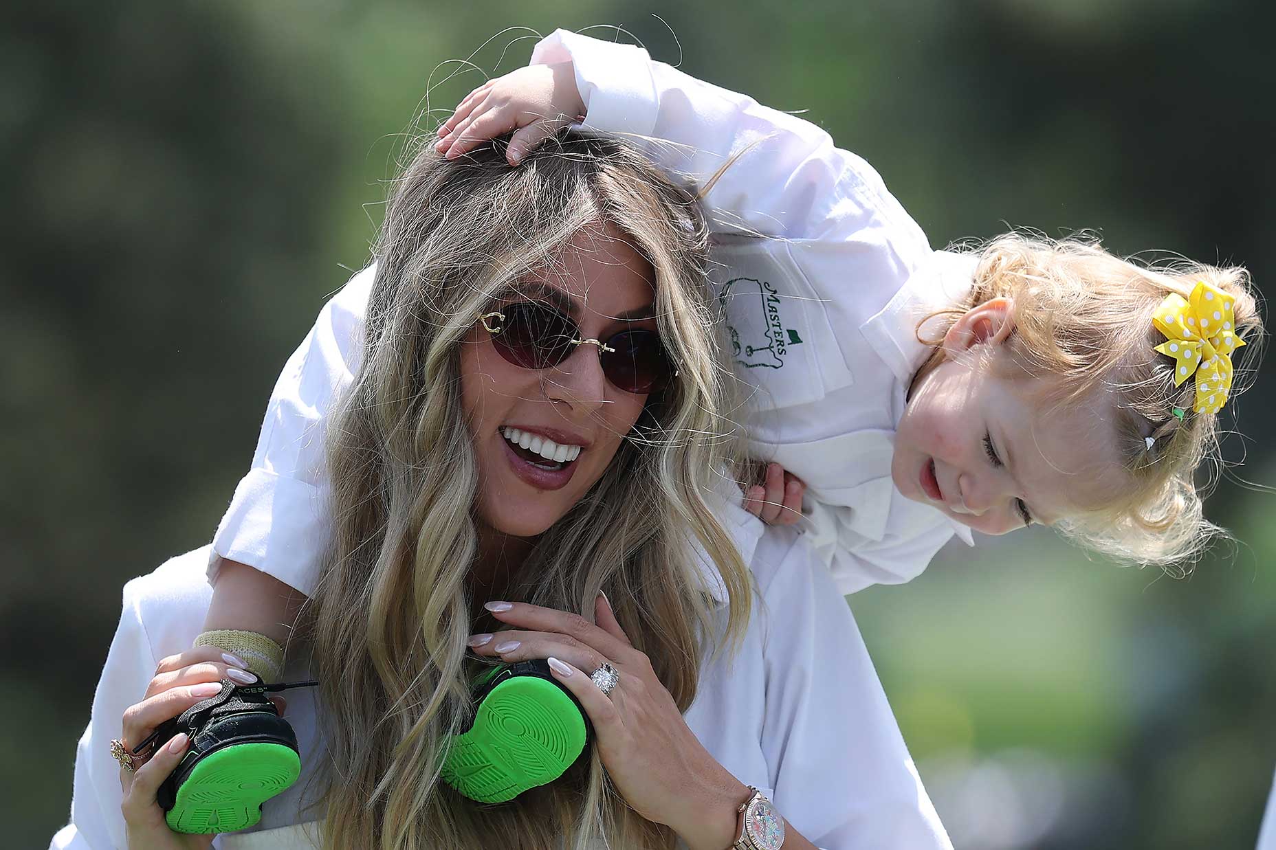 A smiling woman wearing sunglasses carries a young child with blonde hair and a yellow bow on her shoulders outdoors, reminiscent of enjoying a sunny day at the Masters par 3. The child playfully leans over her head; both are dressed in white.