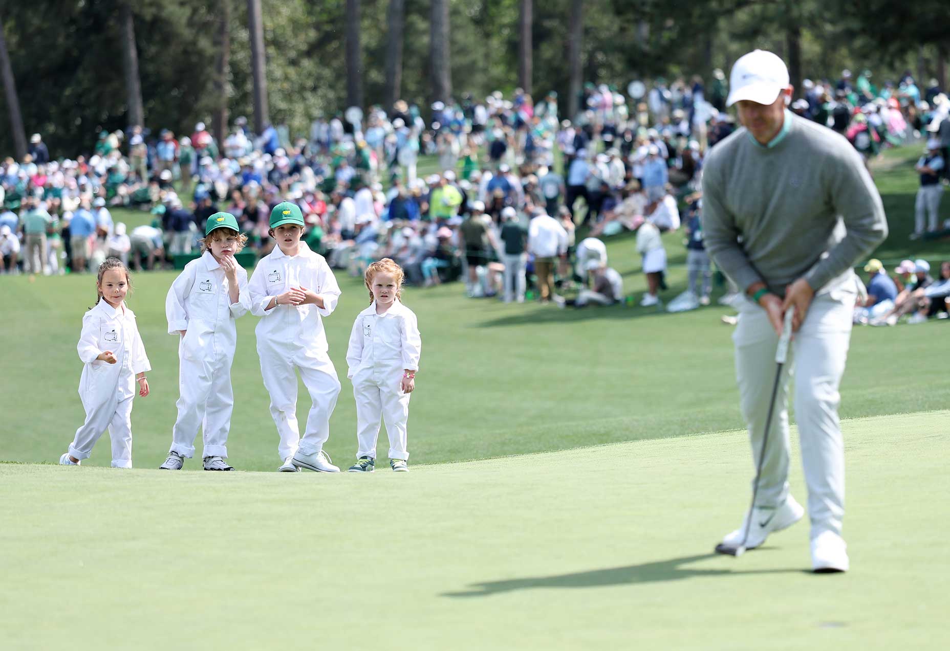 A golfer prepares to putt on a par 3 green at the Masters, while four young children in white jumpsuits and green hats watch nearby. A large crowd sits and stands in the background surrounded by trees.