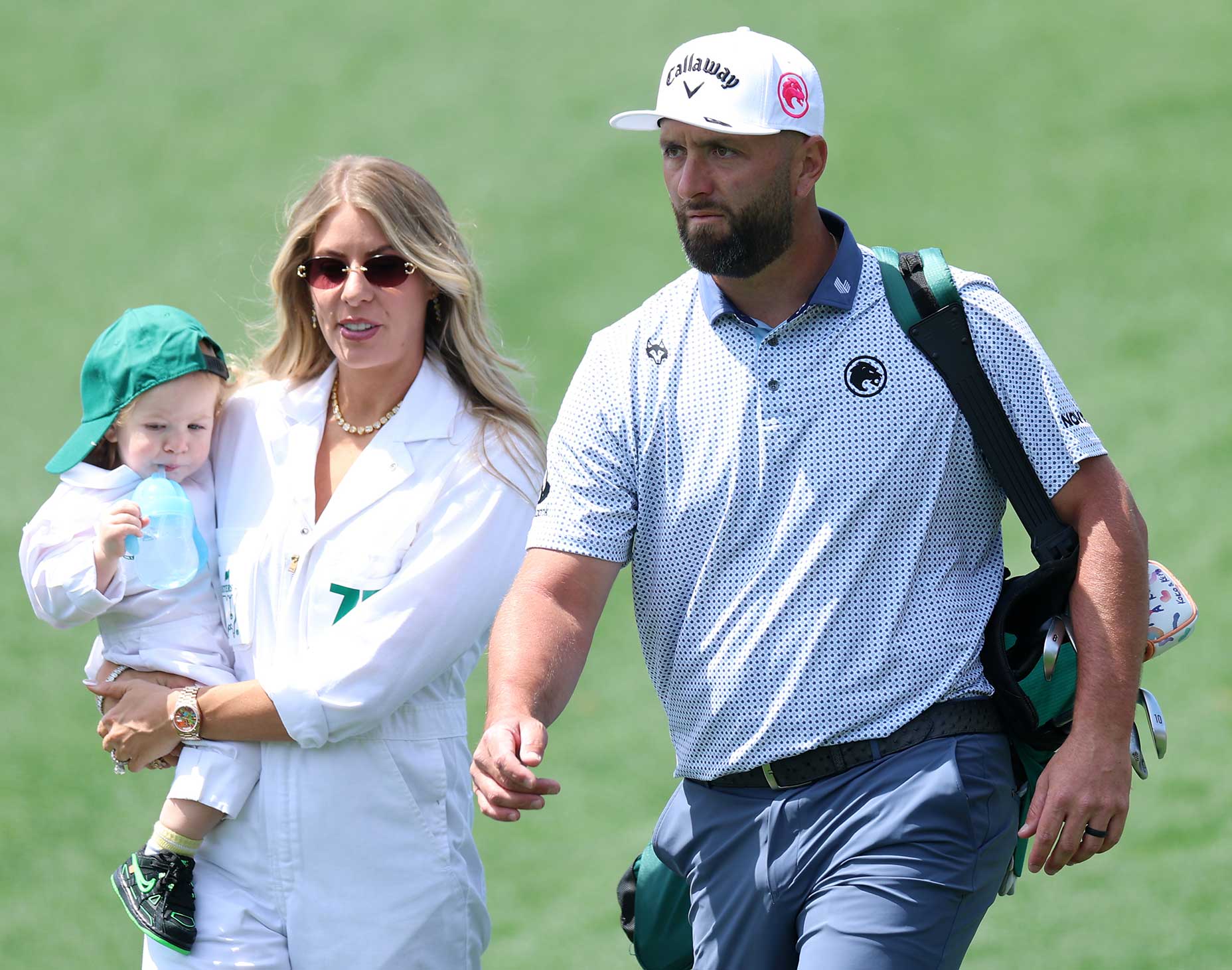 A man in a Masters golf outfit walks on grass with a woman holding a toddler dressed in white and green. The man carries a bag over his shoulder, while the woman and child, both wearing sunglasses, enjoy the par 3 atmosphere together.