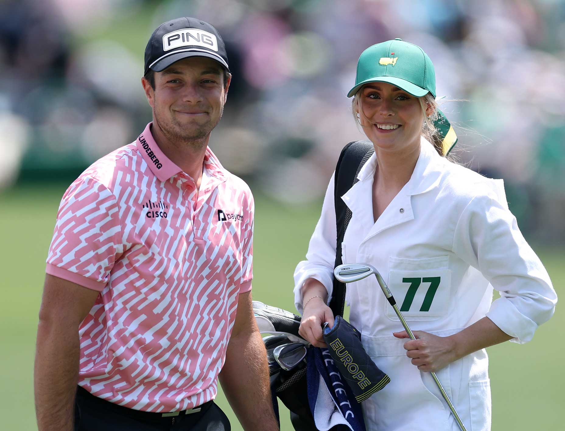 A male golfer in a pink patterned shirt and black cap stands smiling next to a female caddie in a white jumpsuit and green cap, holding golf clubs, on the Masters par 3 course.