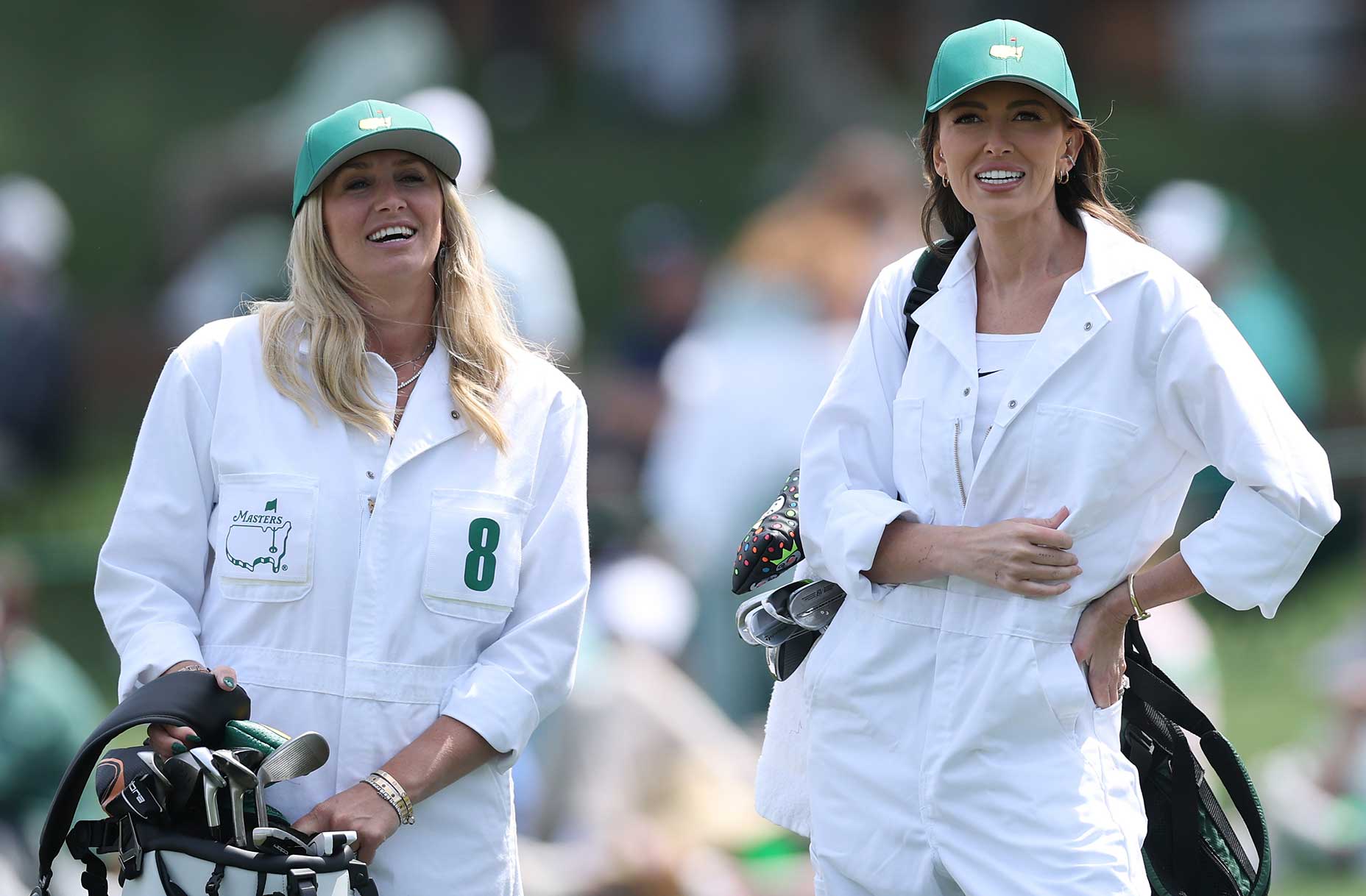 Two women in white coveralls and green hats stand outdoors, smiling. One holds a golf bag, and both sport Masters Tournament logos—capturing the spirit of the par 3 contest amid a blurred backdrop of people and lush greenery.