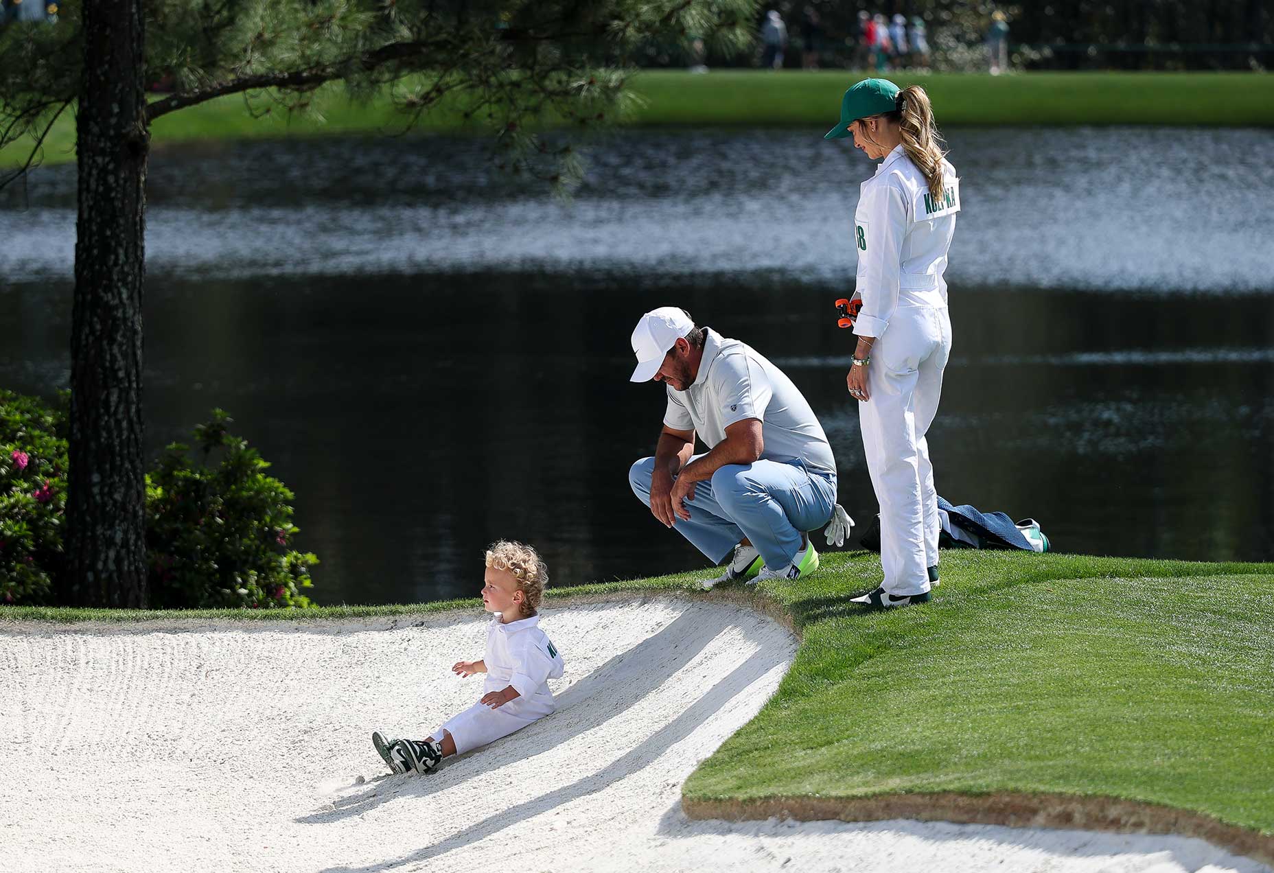 A toddler in a white jumpsuit sits in a sand bunker by a lake on a par 3 hole at the golf course, while a man crouches nearby and a woman in a green hat stands on the grass, watching—the scene recalls Masters traditions.