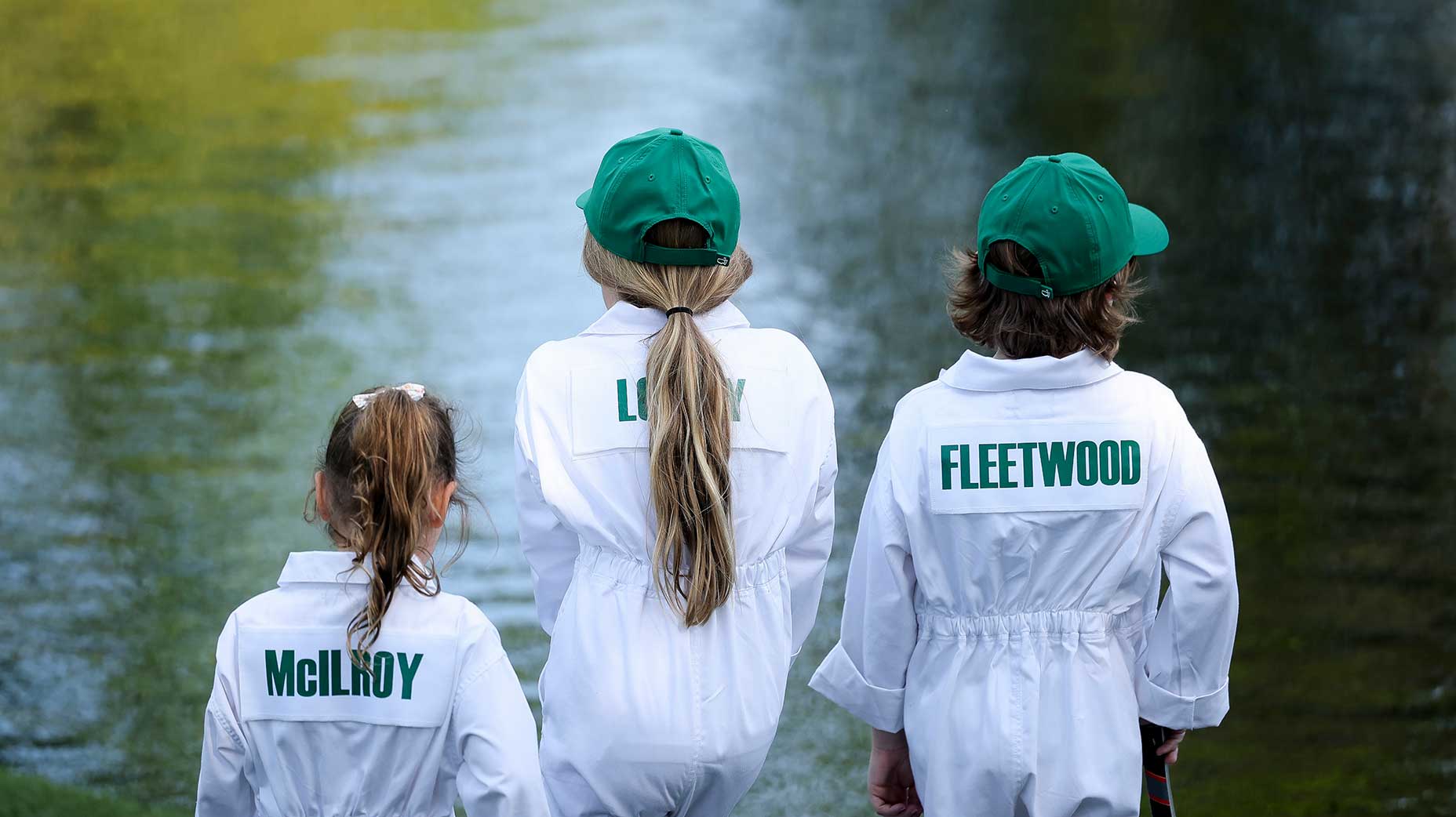 Three children in green caps and white jumpsuits—named McILROY, LOWRY, and FLEETWOOD—stand side by side at the Masters Par 3, gazing out over a serene pond or lake.