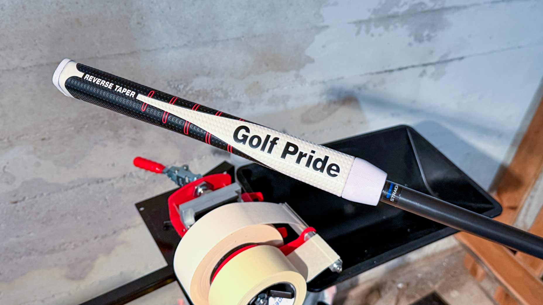 A close-up of a golf club with a white Golf Pride grip and Reverse Taper label, resting on a workbench next to a tape dispenser in a workshop setting.
