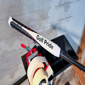 A close-up of a golf club with a white Golf Pride grip and Reverse Taper label, resting on a workbench next to a tape dispenser in a workshop setting.