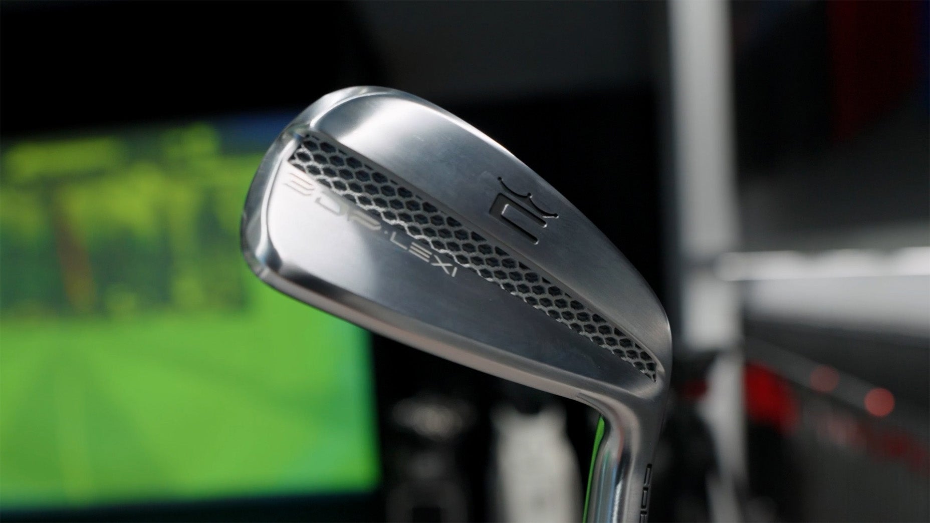A close-up of a silver Cobra 3DP Iron golf club with a hexagonal pattern and the text 20 DYNAMIC engraved on the club head. The background is blurred with shades of green and black.