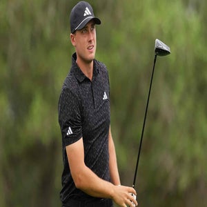 A golfer wearing a black cap and patterned black shirt holds a golf club and looks ahead, with a blurred green background&mdash;perhaps visualizing the 2026 Valero Texas Open Saturday tee times.