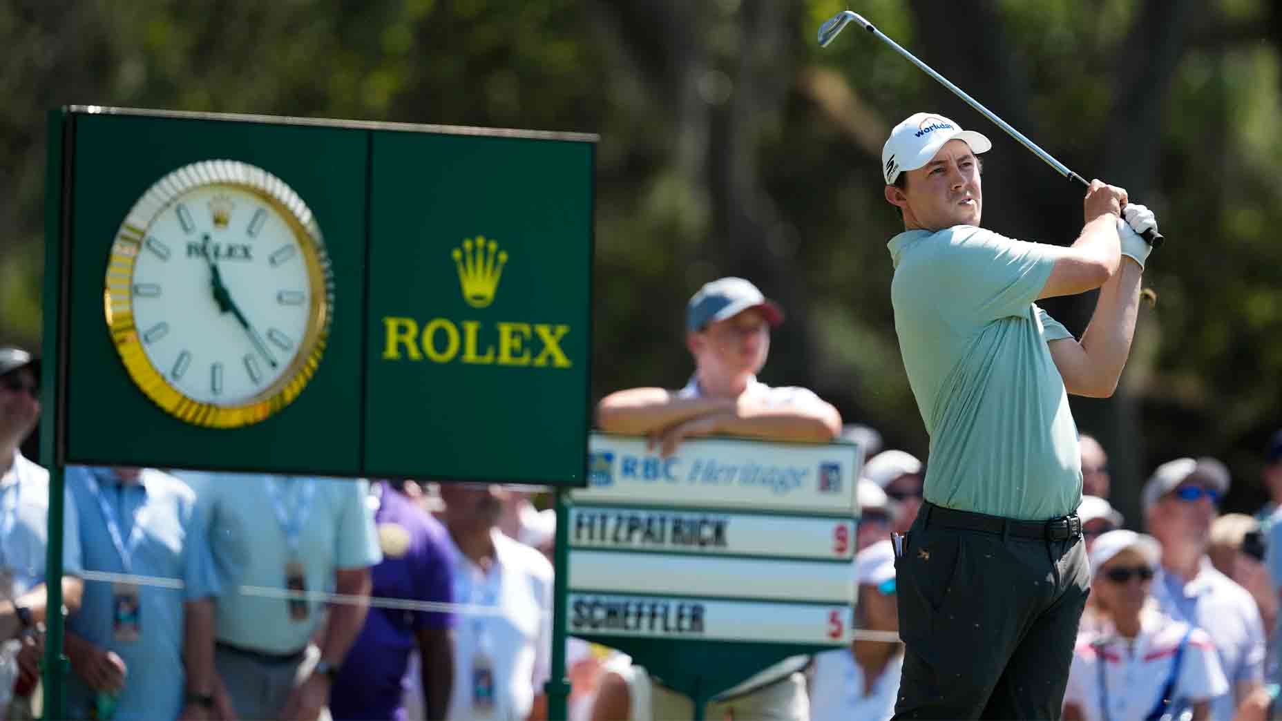 A golfer swings a club during the 2026 RBC Heritage tournament, with a Rolex clock and leaderboard in the background, as a crowd of spectators watches.