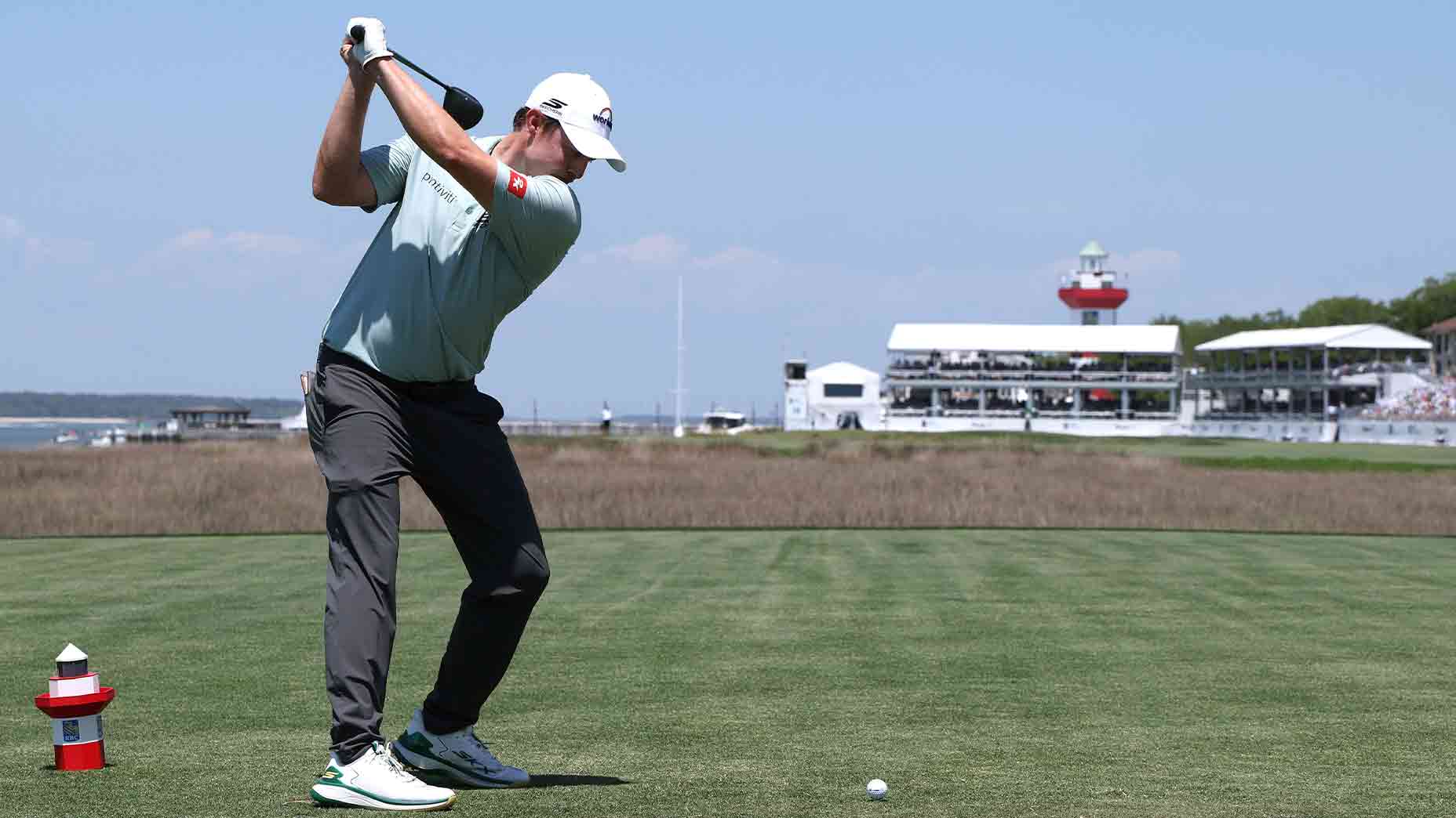 A golfer prepares to swing on a tee box with a golf ball in front of him, as spectators—possibly awaiting the 2026 RBC Heritage Saturday tee times—watch from a white pavilion under a clear blue sky.