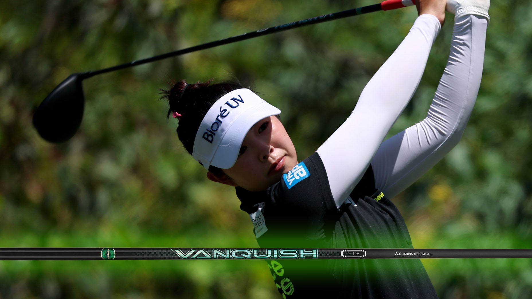 A female golfer wearing a white visor and black shirt follows through on her swing, with a golf club and the word VANQUISH highlighted in the foreground. Lush greenery is blurred in the background.