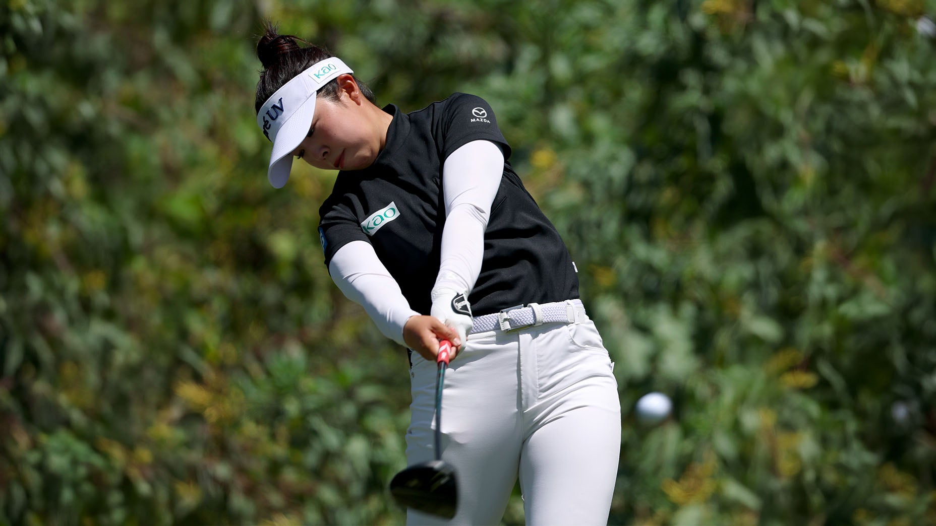 A female golfer in a black shirt and white pants swings a golf club on a lush, green course, her eyes focused on the ball as it leaves the tee.