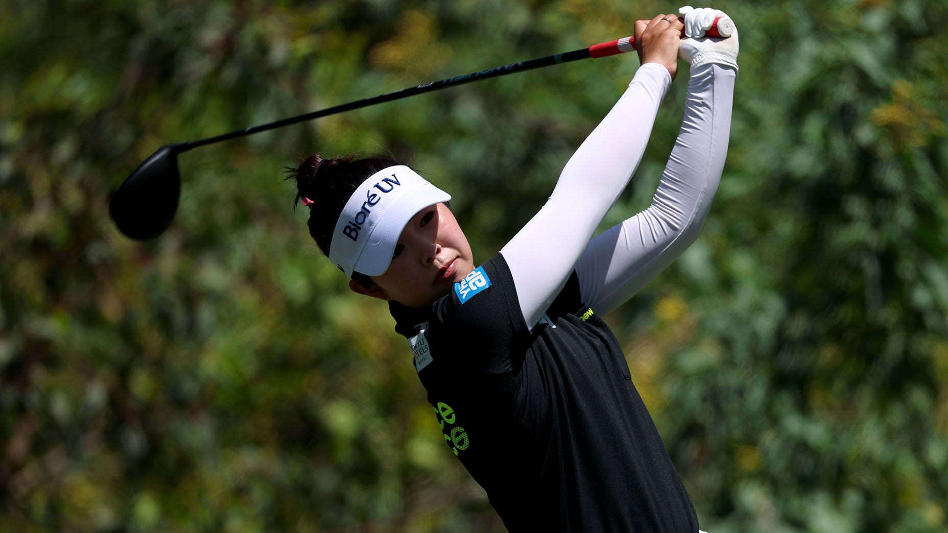 A golfer wearing a white visor, black shirt, and white arm sleeves follows through on a swing with a driver, with greenery visible in the blurred background.