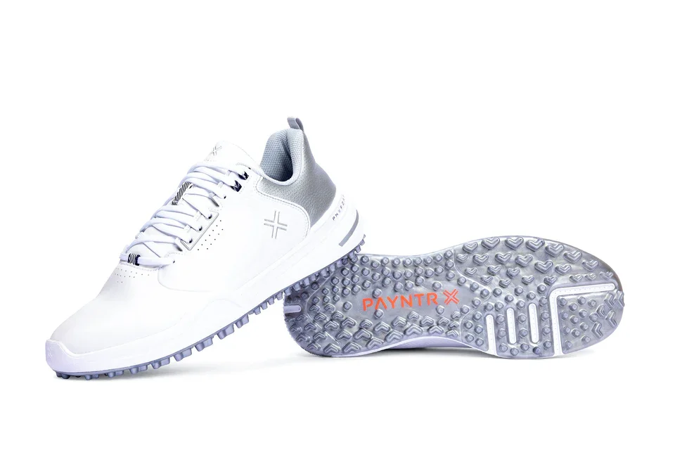 A pair of white athletic golf shoes with gray accents; one shoe is upright, while the other reveals the textured gray sole with PAYNTR X branding in orange&mdash;crafted to be among the best golf shoes 2026 has to offer.