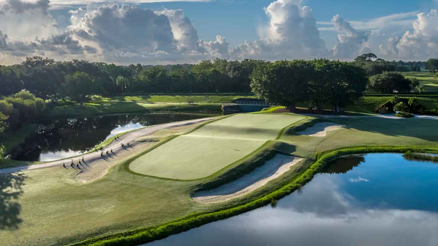A lush golf course in Boca Raton with a putting green surrounded by water hazards, sand bunkers, and trees under a partly cloudy blue sky—perfect for your next golf trip. Golfers are visible walking along the green.