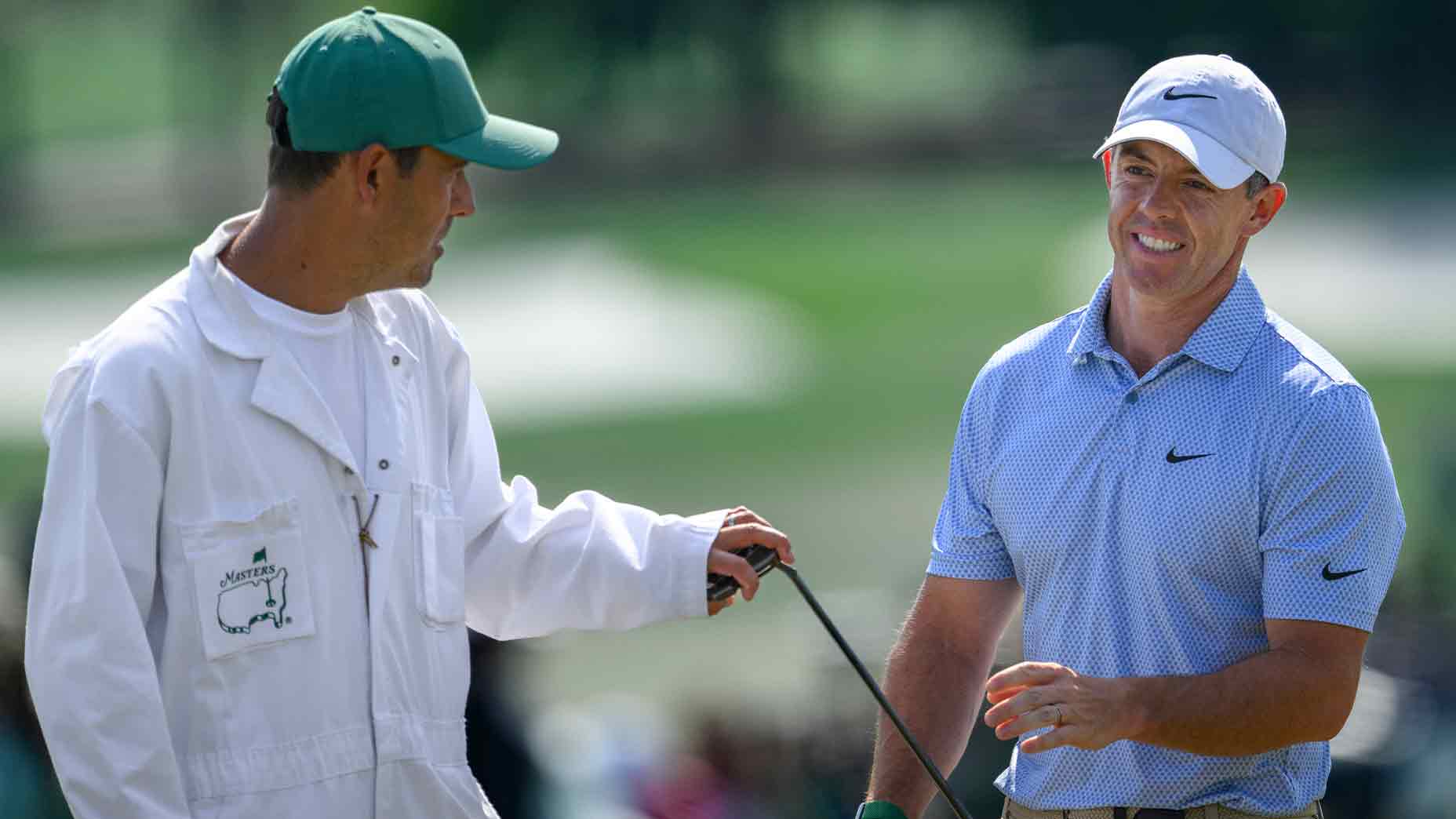 A golfer in a blue shirt and white cap smiles while talking to his caddie, both discussing their next move on the golf course during the Masters, where money and glory are on the line.