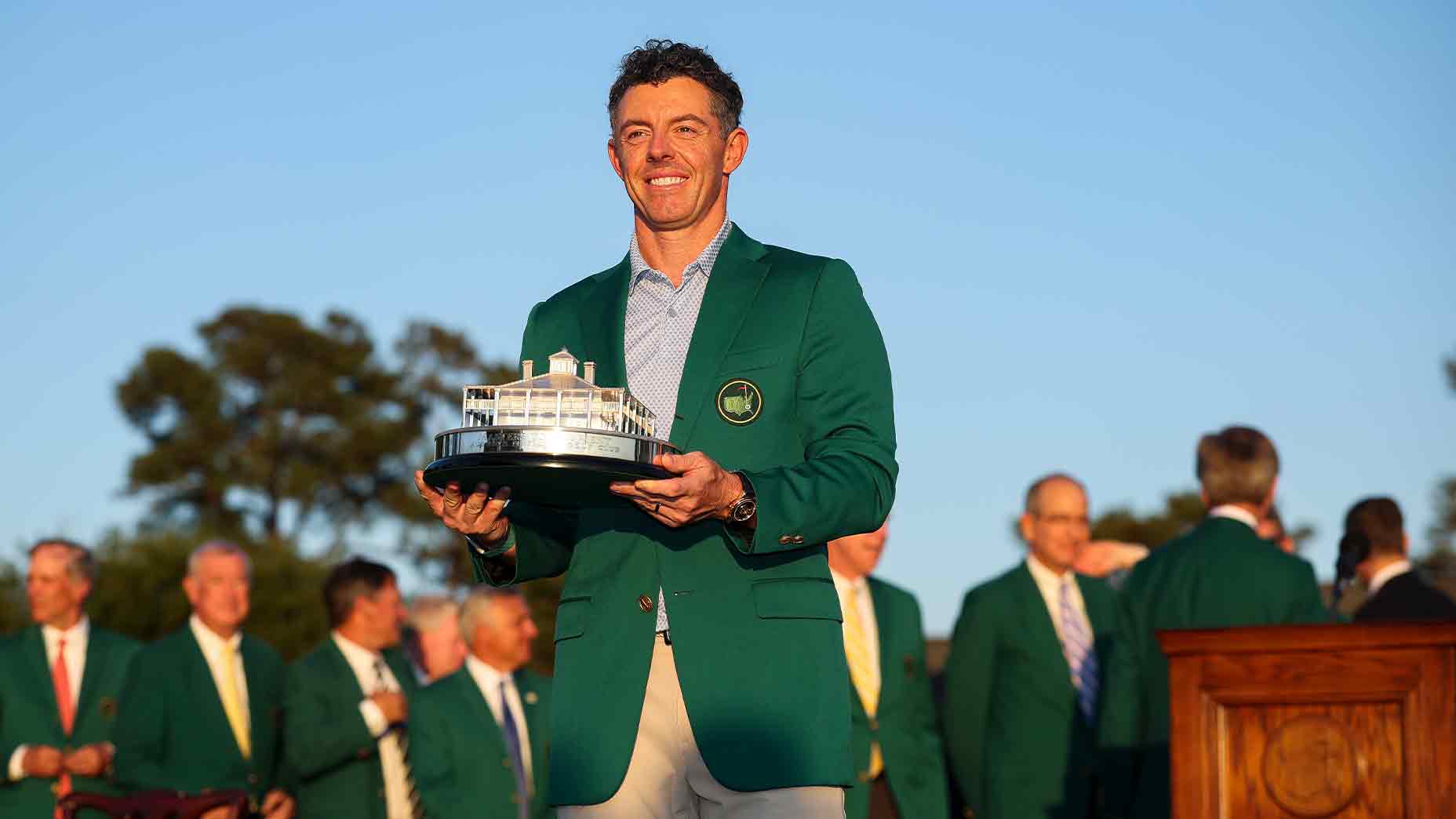 A smiling golfer wearing a green jacket holds a trophy outdoors, celebrating his victory and share of the 2026 Masters purse, with a group of people in suits and green jackets under a clear sky.