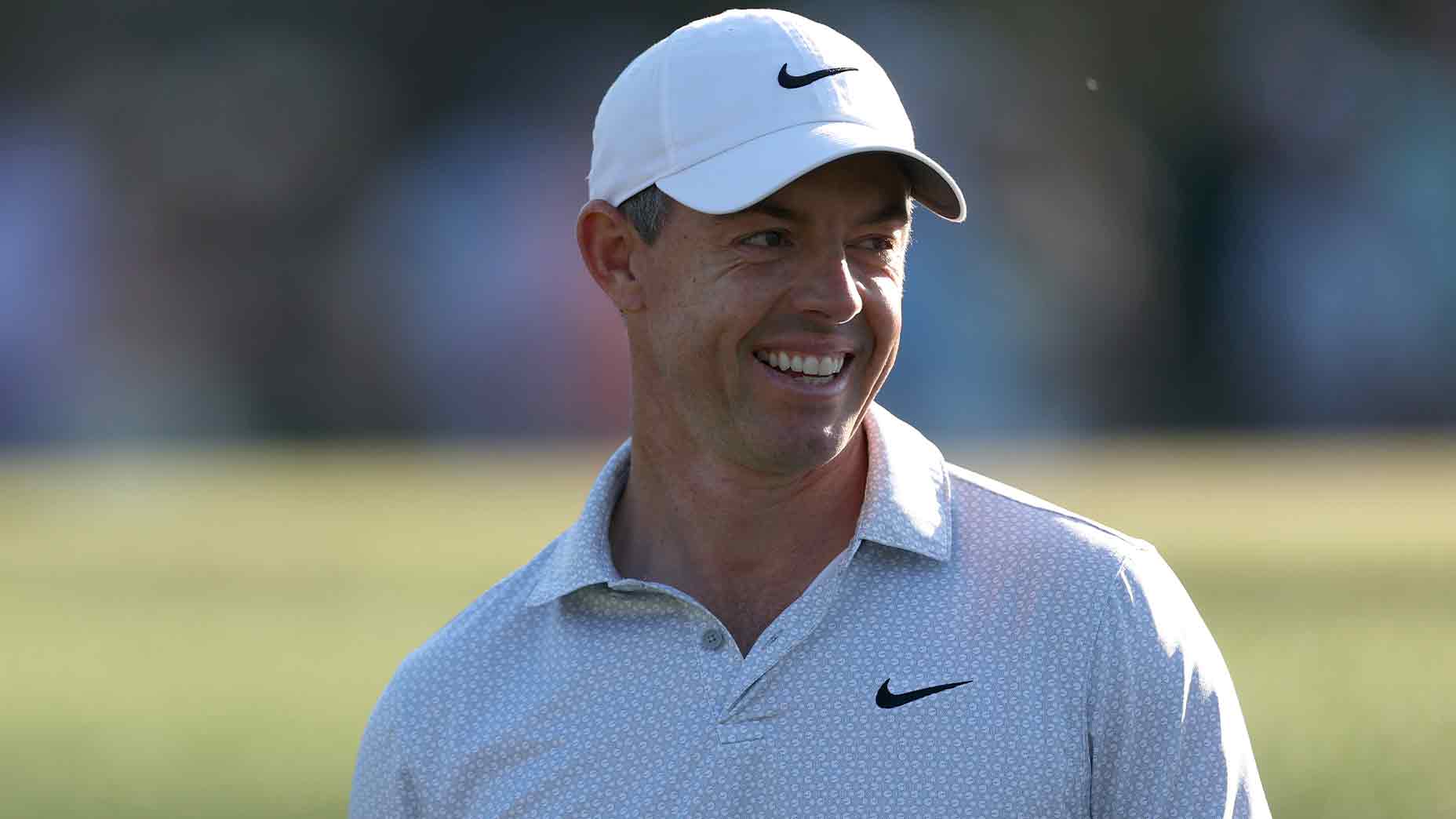 A man wearing a white Nike cap and a light-colored collared shirt with a Nike logo smiles while standing outdoors on a golf course, resembling Rory McIlroy.