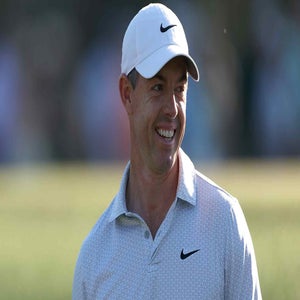 A man wearing a white Nike cap and a light-colored collared shirt with a Nike logo smiles while standing outdoors on a golf course, resembling Rory McIlroy.