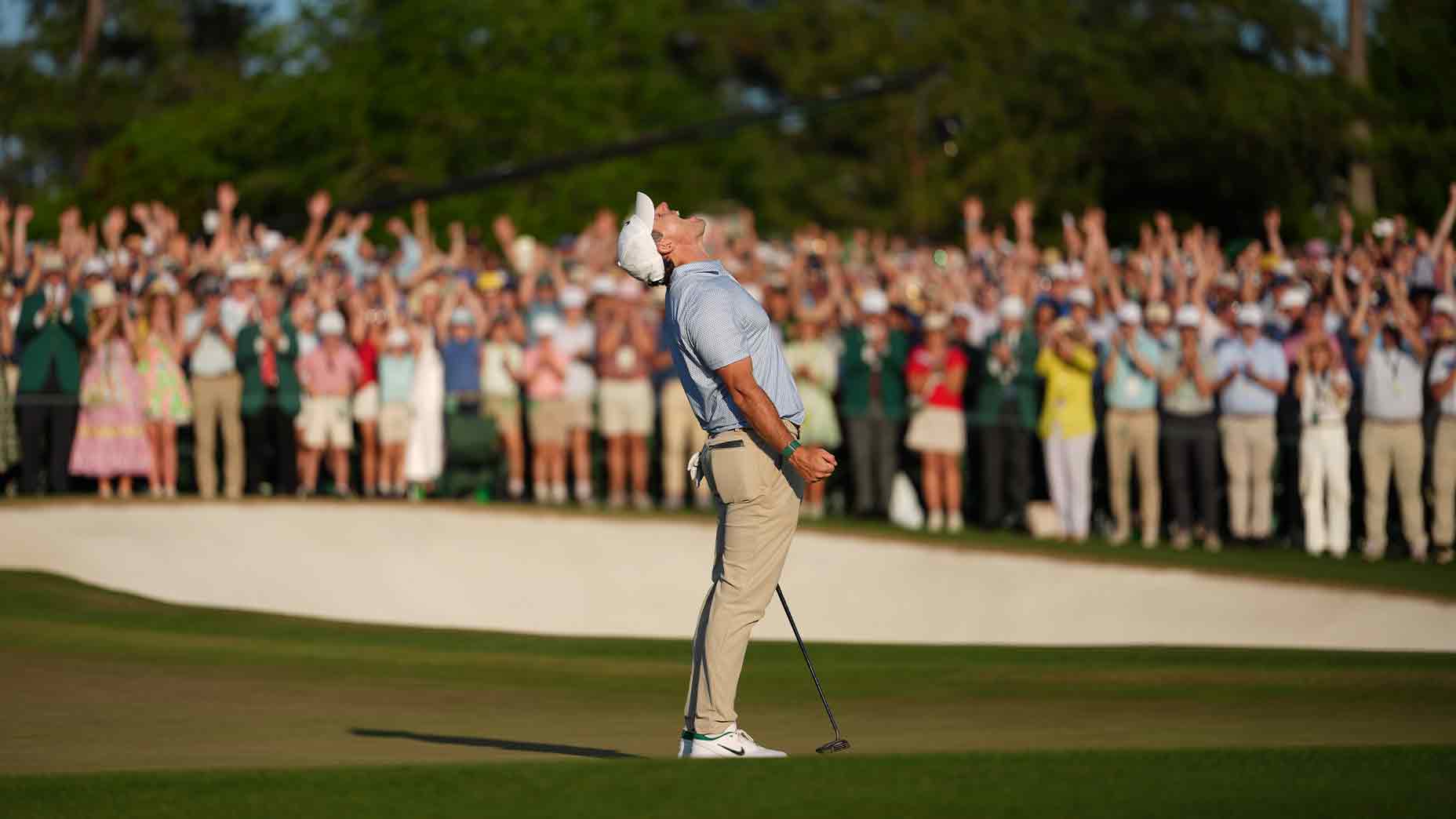 A golfer stands with his head tilted back and arms slightly raised in celebration on the Masters green, while a large, cheering crowd applauds in the background.