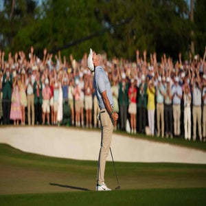 A golfer stands with his head tilted back and arms slightly raised in celebration on the Masters green, while a large, cheering crowd applauds in the background.