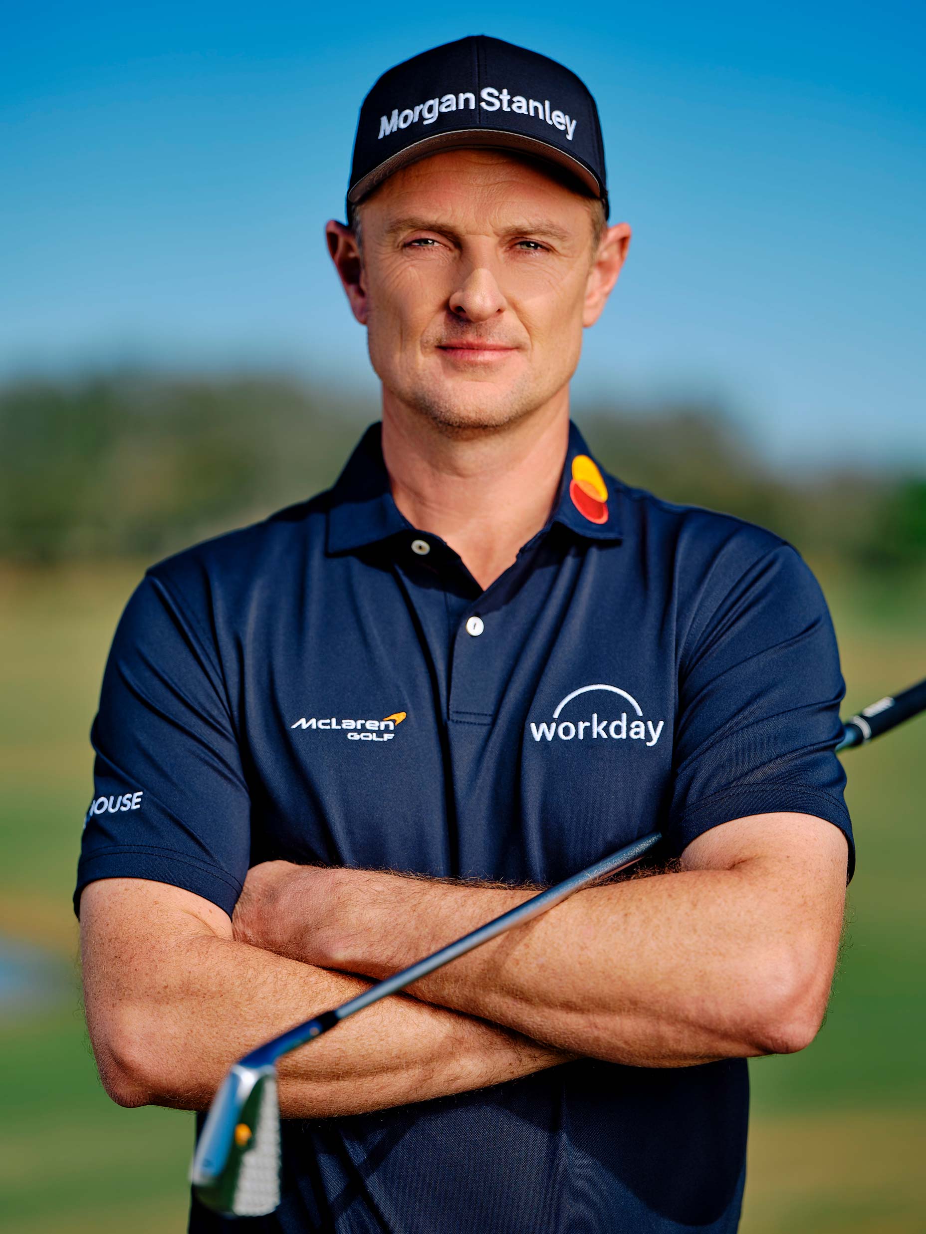 A male golfer, Justin Rose, stands outdoors with arms crossed, wearing a navy blue polo shirt and a cap branded with Morgan Stanley and Workday. He holds a golf club, with a golf course and blue sky in the background—a scene after his shocking move to McLaren.