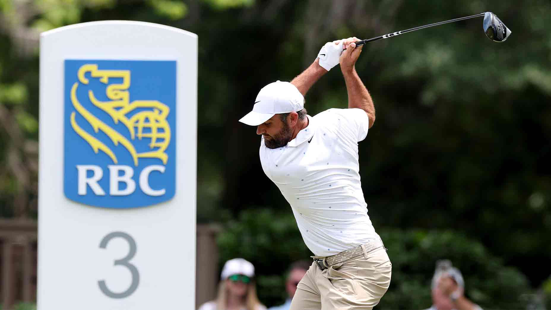 A golfer in a white shirt and cap swings a club in front of a large sign displaying the RBC logo and the number 3, with greenery and spectators blurred in the background, reminiscent of scenes from tournaments like those with Valero Texas Open purse stakes.