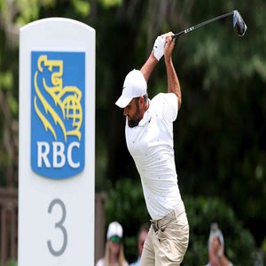 A golfer in a white shirt and cap swings a club in front of a large sign displaying the RBC logo and the number 3, with greenery and spectators blurred in the background, reminiscent of scenes from tournaments like those with Valero Texas Open purse stakes.