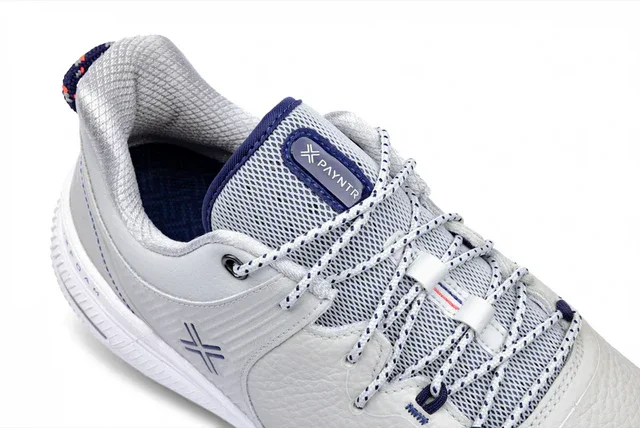 A close-up of a white sneaker with blue and gray accents, featuring a textured upper, patterned laces, and the PAYNTR logo. With its cushioned collar and detailed sole design, it's set to be among the best golf shoes 2026 has to offer.