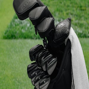 A close-up of a golf bag filled with various golf clubs covered in black headcovers, positioned on a grassy area. A white towel hangs from the side of the bag.
