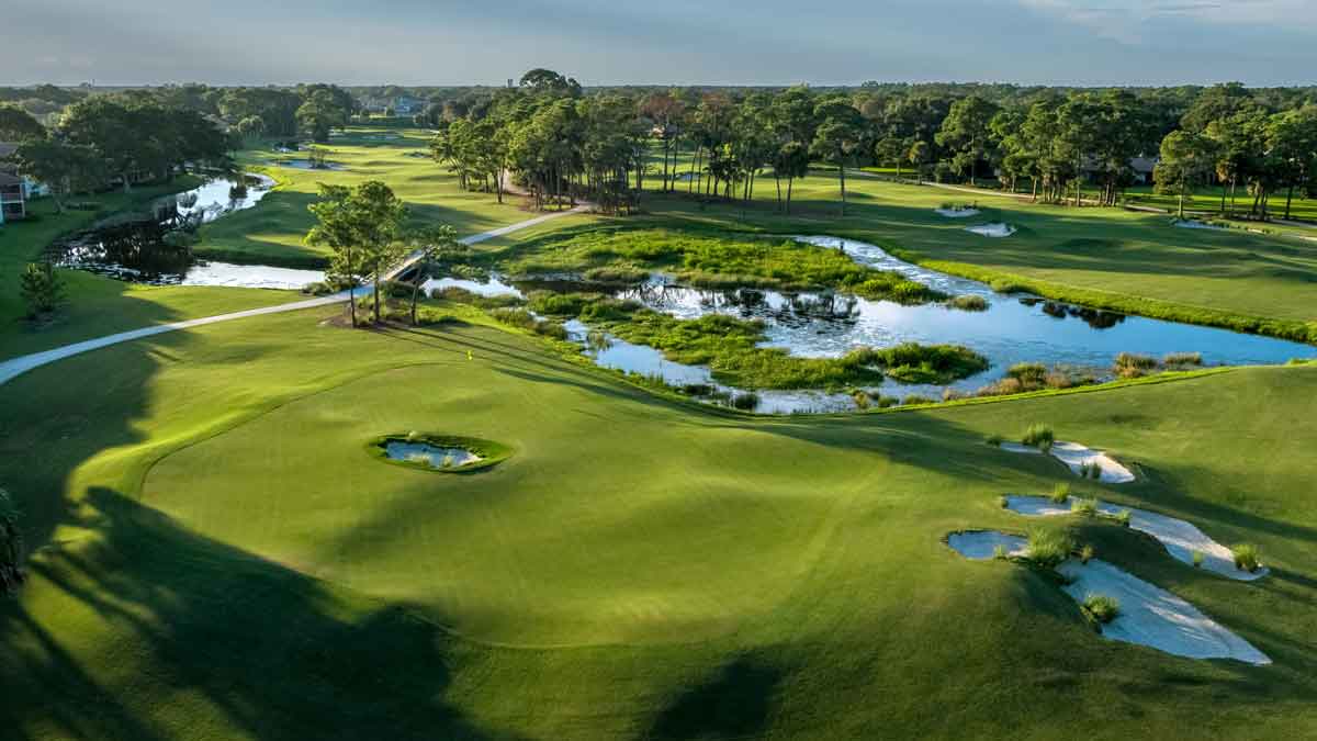 Aerial view of a lush Boca Raton golf course featuring manicured greens, sand bunkers, winding pathways, water hazards, and surrounding trees under a clear sky—the perfect setting for your next golf trip.
