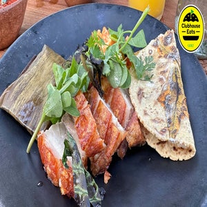 A black plate with crispy pork belly slices, a folded tortilla, fresh greens, and a tamale wrapped in a banana leaf offers exotic bites, with a yellow Clubhouse Eats sticker in the top right corner.