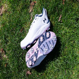 A pair of white Under Armour Drive Pro Clone LE golf shoes, one upright and one showing the sole with multiple cleats, rests on green grass in bright sunlight.
