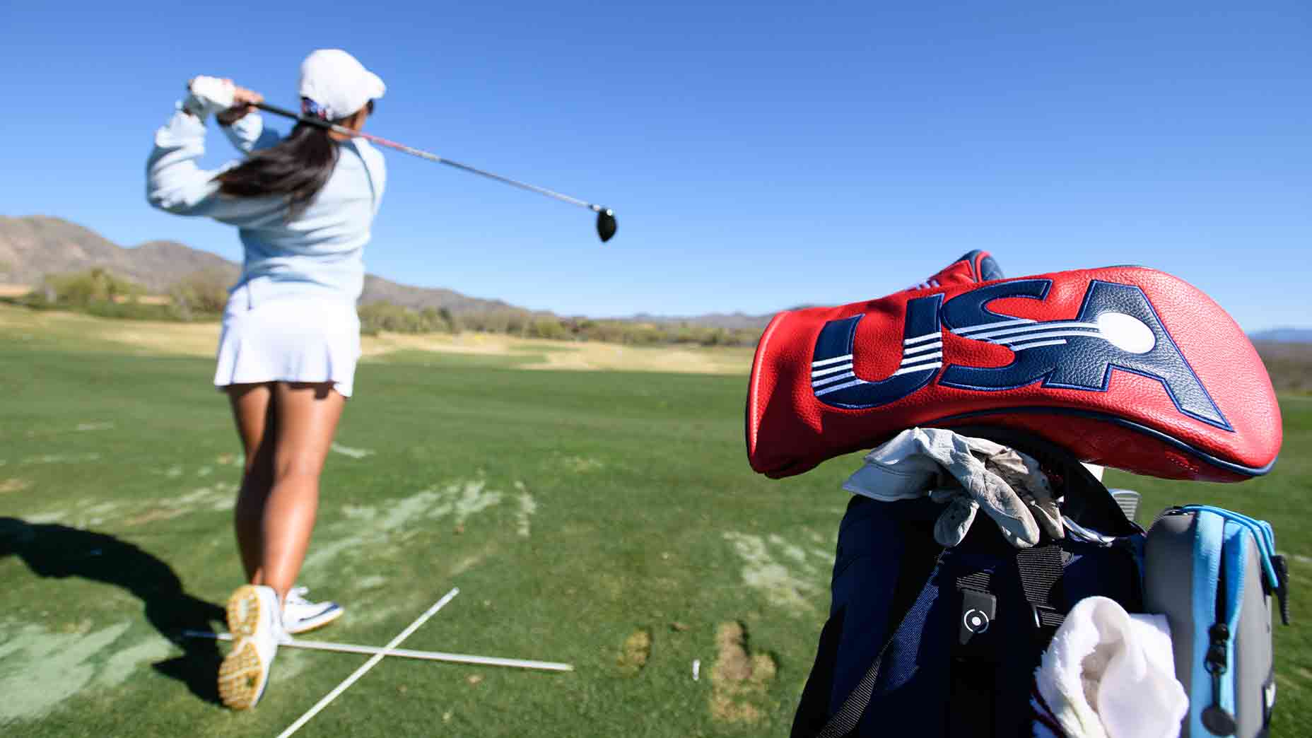 A golfer in a white skirt and cap swings a club on a green course. In the foreground, a golf bag with a red USA headcover hints at U.S. National Junior Team pride. Blue sky and mountains inspire every practice swing.