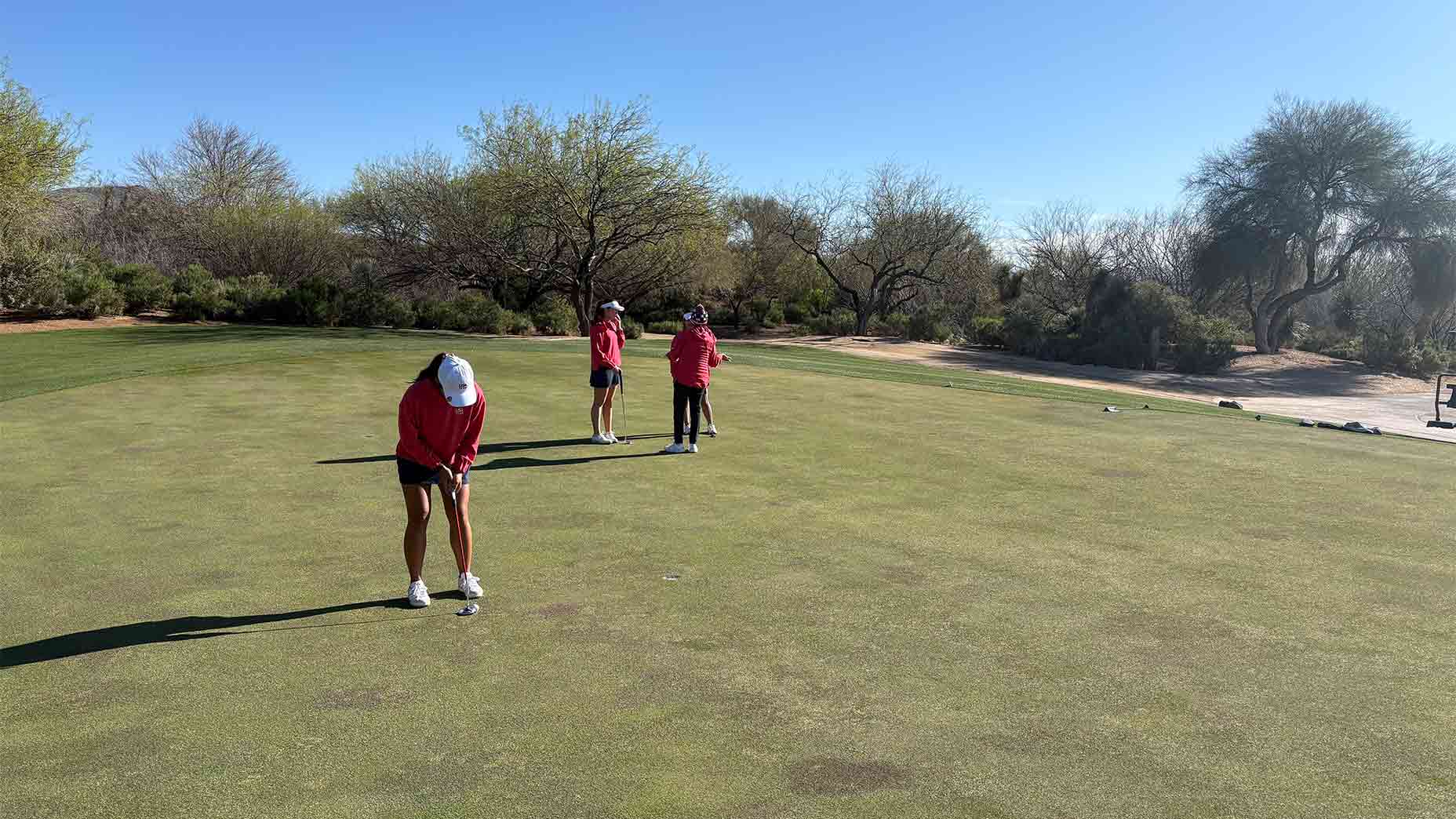 Three people in red shirts and hats are on a golf green, practicing as part of a junior team training session. One putts while the others watch, with trees, bushes, and a clear blue sky in the background.