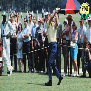 A golfer in a yellow shirt swings his club as a crowd of spectators watches. Some people stand behind a rope, with one holding a red and black umbrella. The scene appears to be at a professional golf tournament.