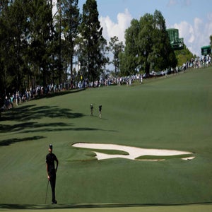 A golfer stands on a green beside a sand bunker on a large, sloping golf course with trees and crowds of spectators lining the fairway in the background.