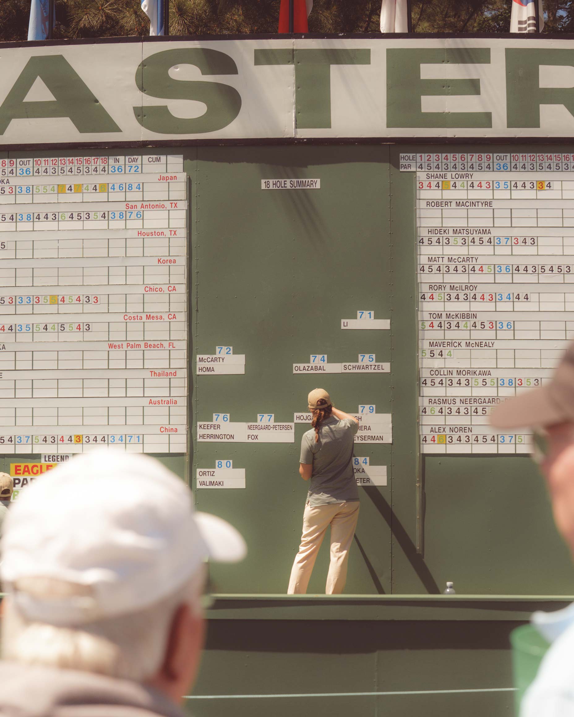 A person updates scores on a large, outdoor Masters golf tournament leaderboard at Augusta National while spectators watch from the foreground. Various player names and numbers are displayed on the board.