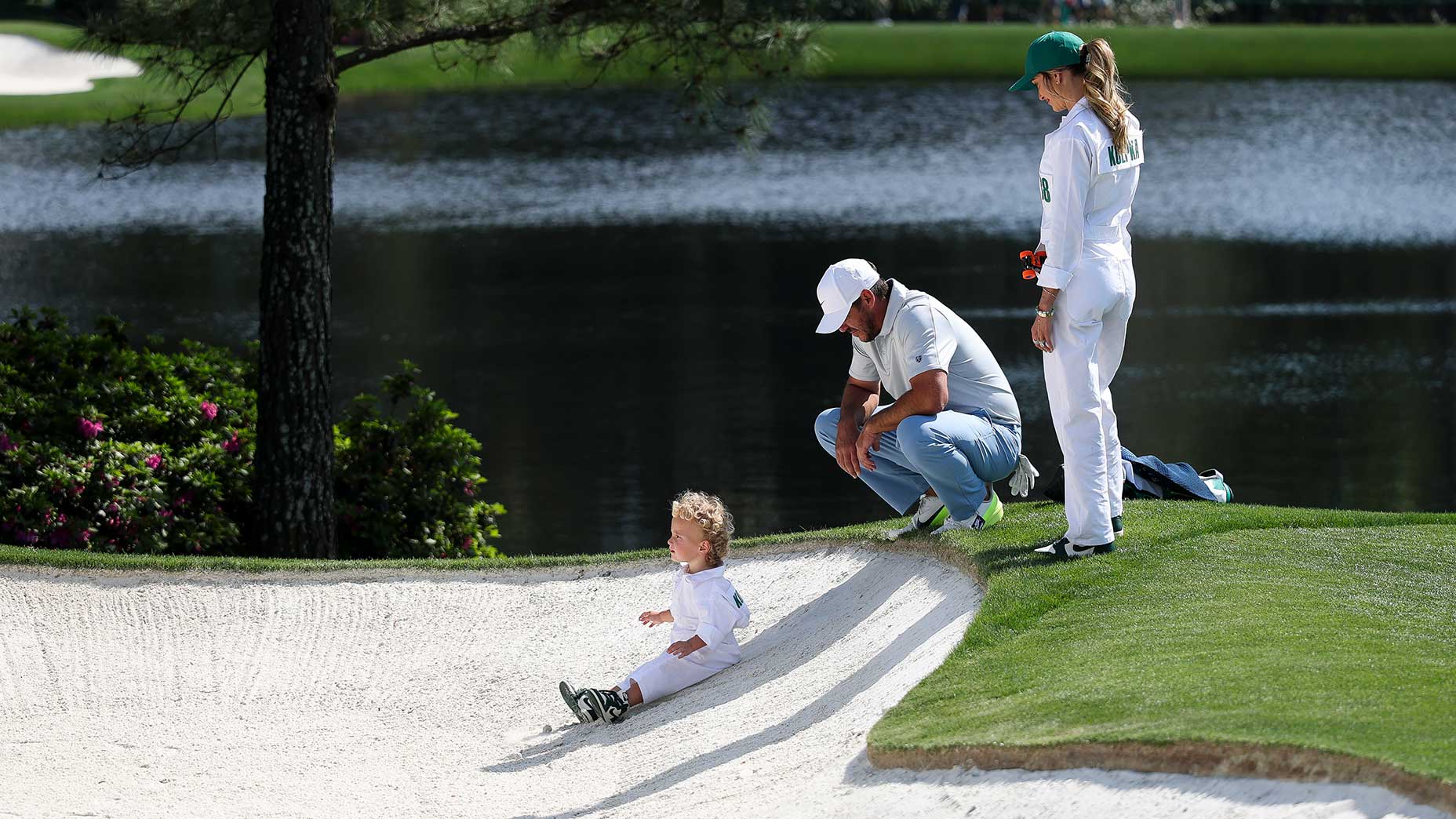 A small child in a white jumpsuit sits in a sand trap on a golf course, while two adults, also in white, watch nearby. A pond and greenery are visible in the background.