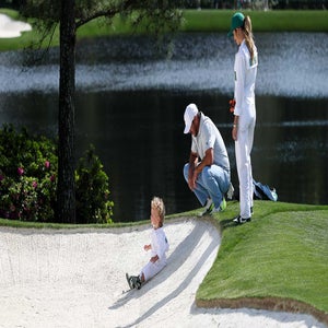 A small child in a white jumpsuit sits in a sand trap on a golf course, while two adults, also in white, watch nearby. A pond and greenery are visible in the background.