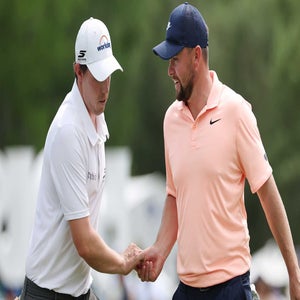 Two male golfers shake hands on a golf course, perhaps discussing the Zurich Classic purse or winner's share. One wears a white shirt and cap; the other, a light peach polo and navy cap. Focused expressions stand out against lush greenery and blurred spectators.