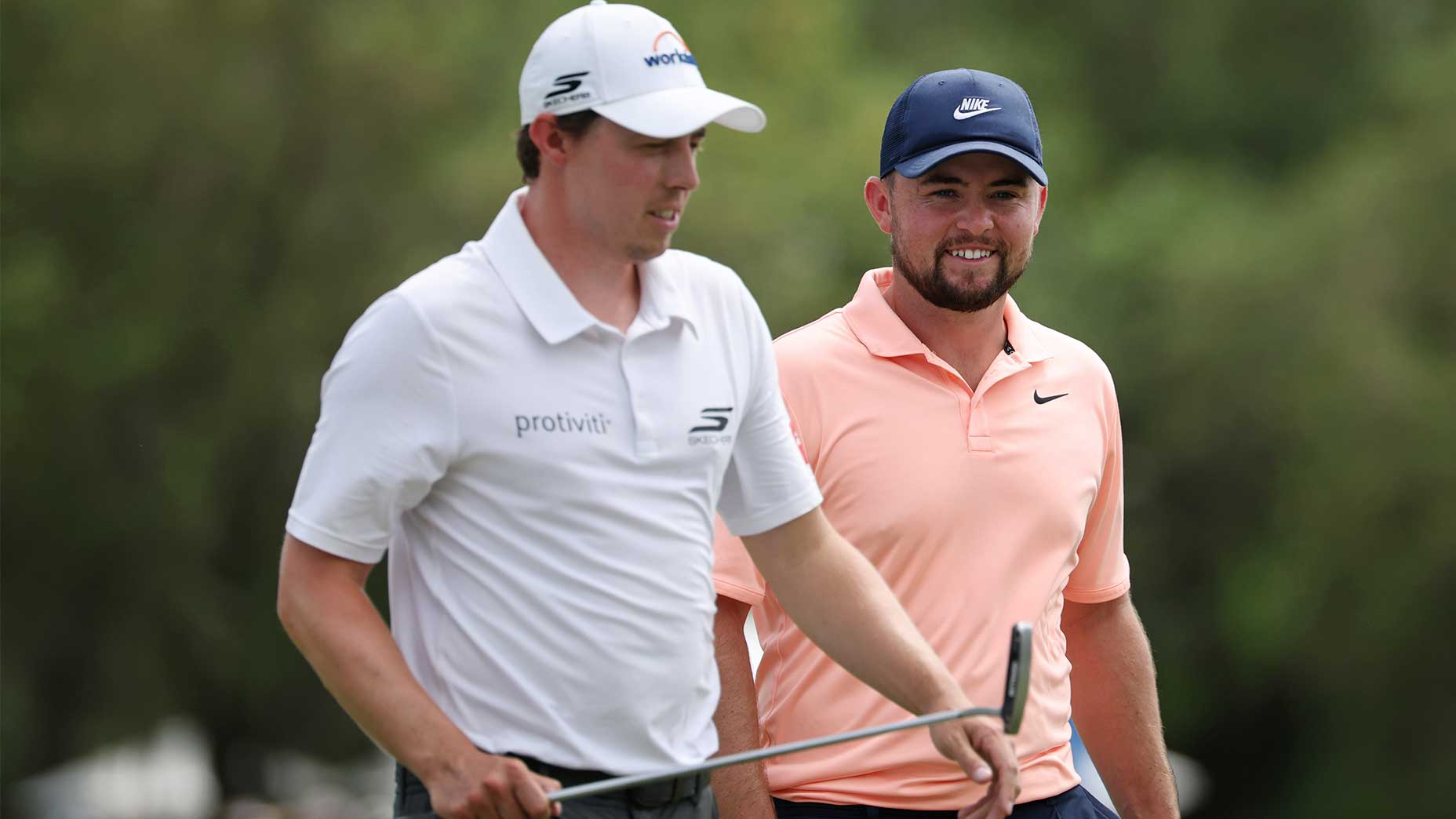 Two male golfers walk on a golf course, smiling as they chat about the 2026 Chevron Championship. One wears a white cap and shirt and holds a putter, while the other sports a peach polo and navy cap, with trees blurred in the background.