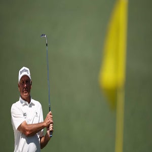 Fred Couples wearing a white shirt and cap holds a golf club at the Masters, with a blurry yellow flag visible in the foreground.