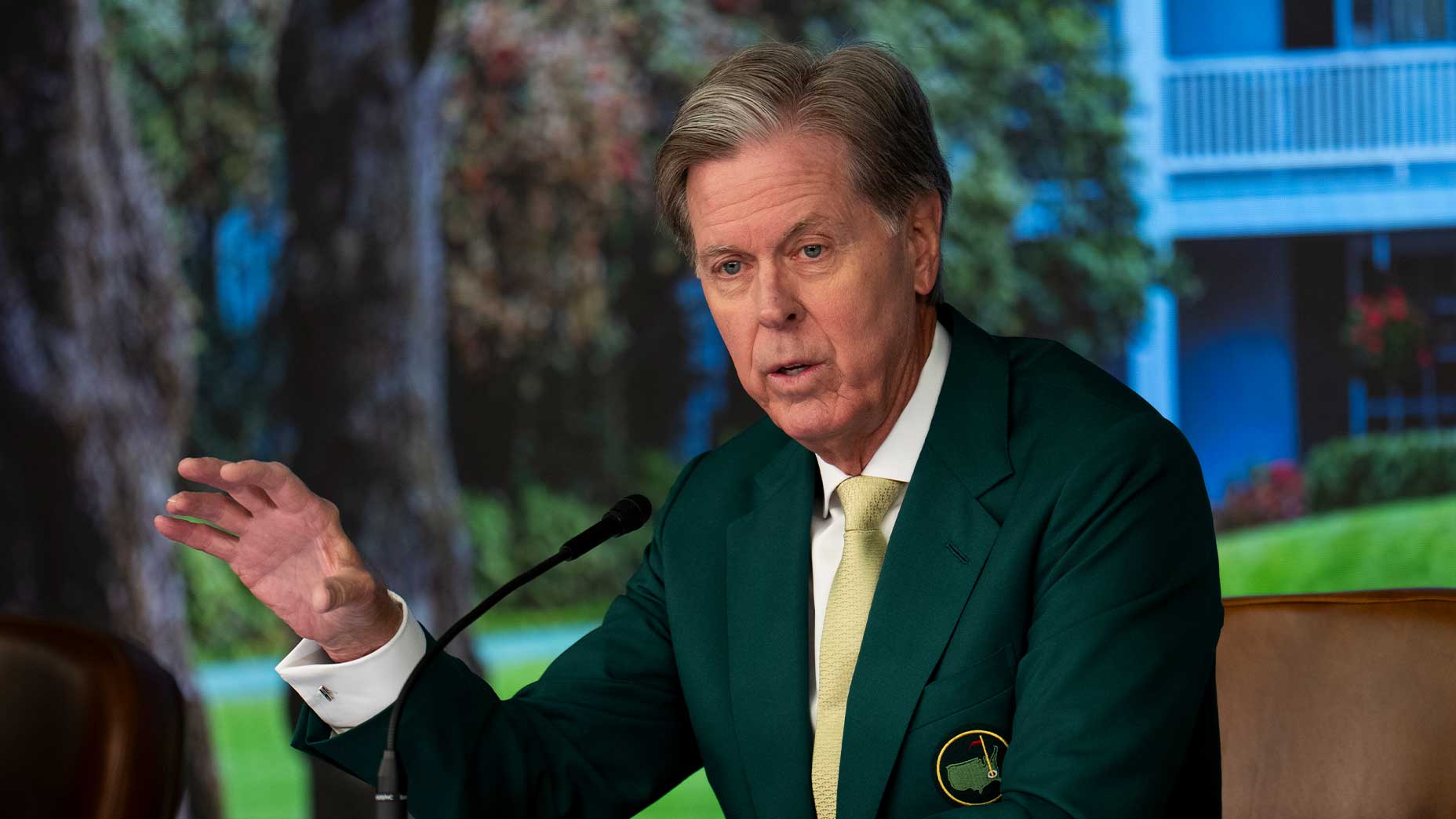 Fred Ridley wearing a green jacket and yellow tie speaks into a microphone at a press conference, gesturing with one hand. A blurred outdoor scene with trees and a building is in the background.