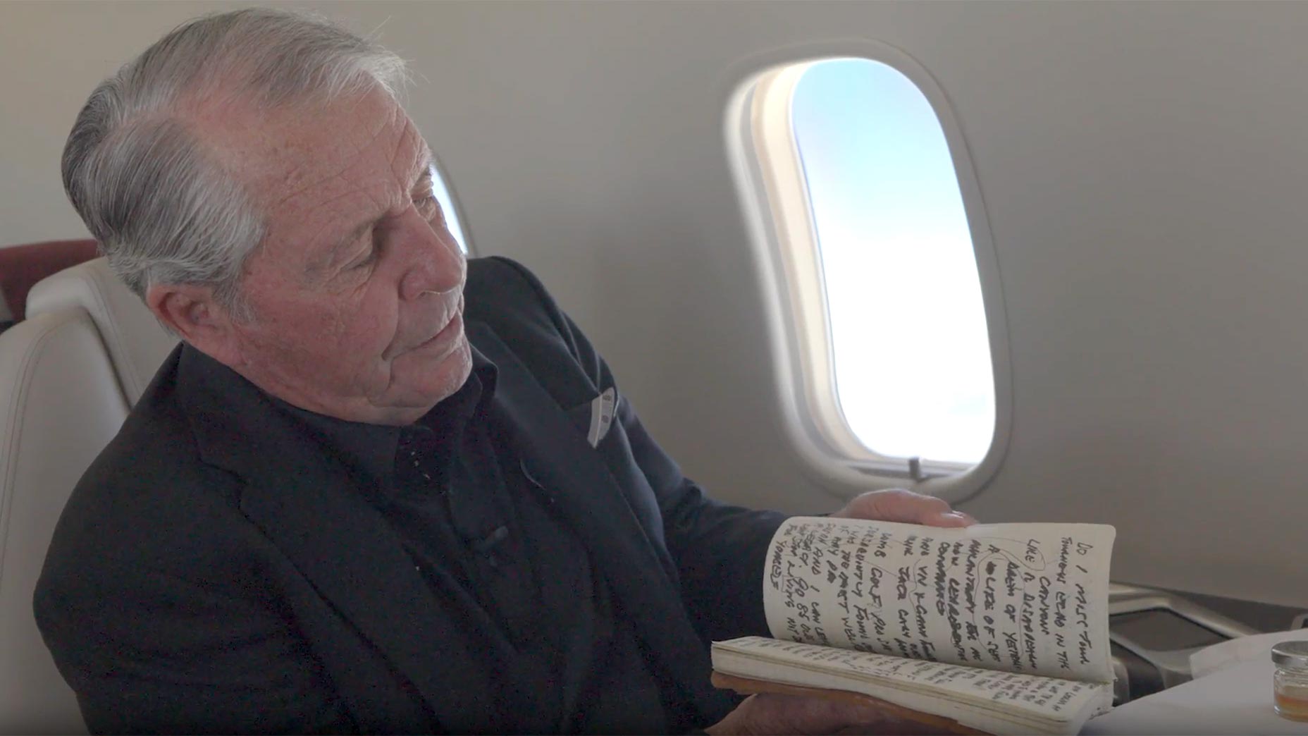 An older man in a dark suit sits on an airplane, reading a handwritten journal. As he flew to the Masters, daylight streams through the window beside him and a drink rests on the tray table, evoking memories of Gary Player’s legendary journeys.